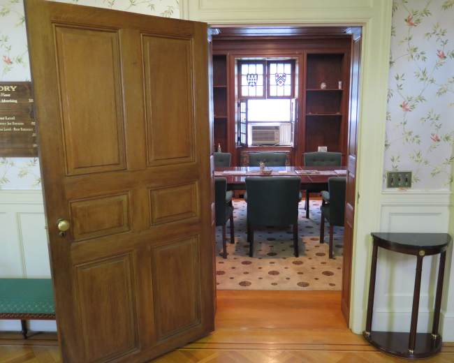 A wooden door opens to a meeting room with a long table surrounded by chairs, located in a space with floral wallpaper and a window featuring an air conditioning unit.