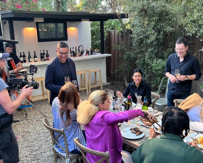 A group of people is gathered around a table in an outdoor setting, sharing food and drinks while a camera crew films the scene.