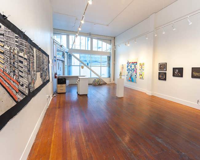 The image shows an art gallery featuring a variety of artworks displayed on white walls and wooden flooring, with natural light coming in through large windows.