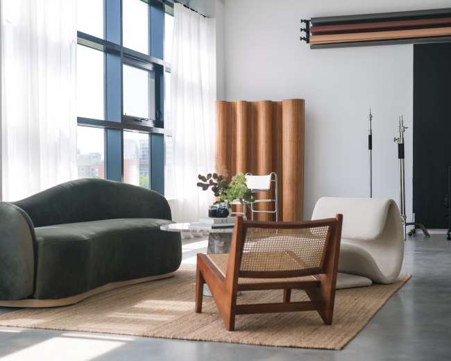 The image shows a modern, well-lit living space featuring a green sofa, a white lounge chair, and a wooden accent chair on a textured rug.