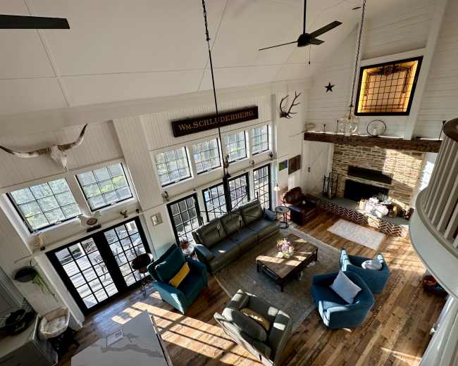 The image shows a spacious living room with large windows, a stone fireplace, and a mix of modern and rustic furniture.