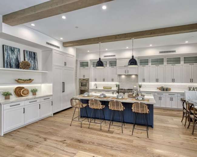 The image shows a modern kitchen with white cabinetry, a blue island, and wooden beams overhead, featuring barstools and contemporary lighting.