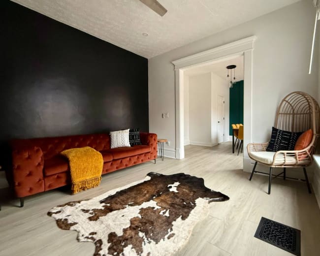 The image shows a spacious living room featuring a brown tufted sofa, a cowhide rug on the floor, and a stylish woven chair beside a dark accent wall.