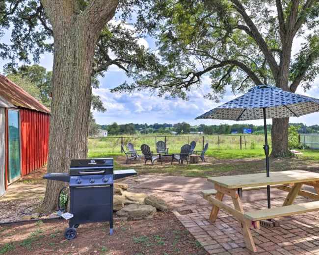 A backyard with a barbecue grill, a picnic table with an umbrella, and seating arranged around a fire pit, set against a backdrop of greenery and a fence.