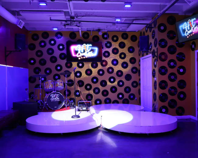 The image shows a stage area featuring a microphone stand, a drum set, and wall decor made of black vinyl records, illuminated by purple lighting.