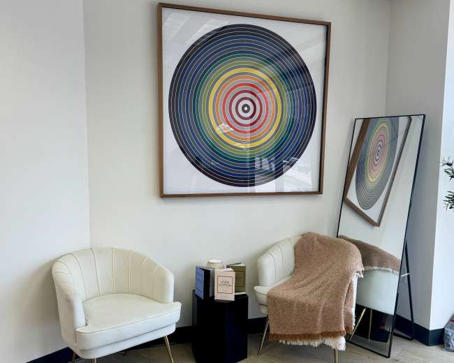 The image shows a corner of an office featuring two white chairs, a small side table, a colorful circular artwork on the wall, and a large mirror.