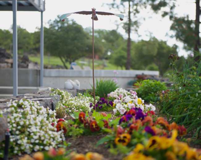 A garden with colorful flowers and a bird sculpture stands in the foreground, while a stone wall and green hillside are visible in the background.