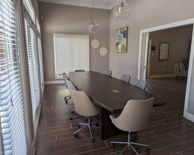 A long, dark conference table with six gray chairs is positioned in a well-lit meeting room featuring large windows and wood-like flooring.
