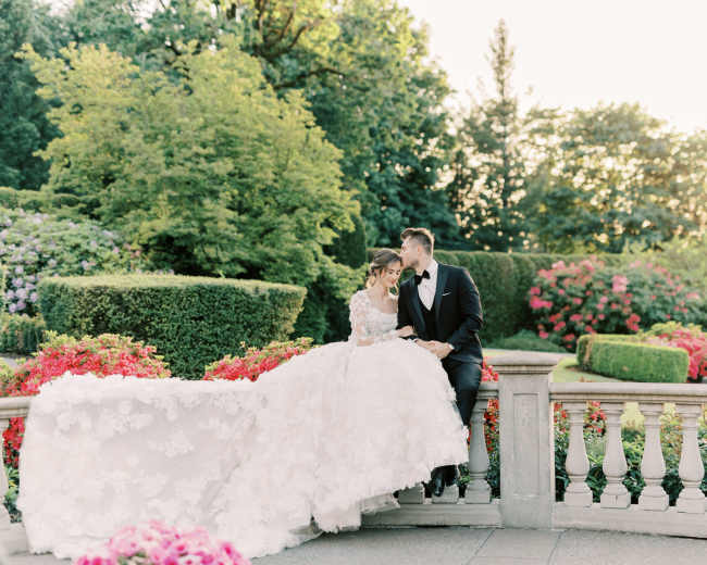 A couple in formal attire sits on a stone railing surrounded by lush gardens and vibrant floral arrangements.