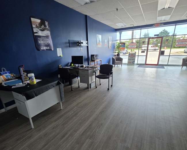 The image shows the interior of a business space with a reception area, including a desk, chairs, and large windows allowing natural light to enter.