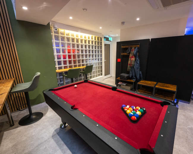 The image shows a modern recreational room featuring a red felt pool table, surrounded by seating areas and a coat rack in a well-lit space.