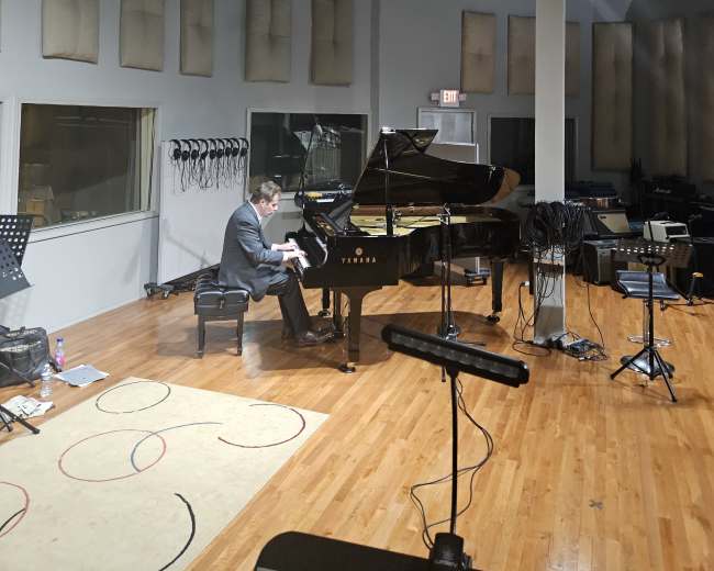 A musician is playing a grand piano in a sound studio with acoustic panels on the walls and various music equipment in the background.