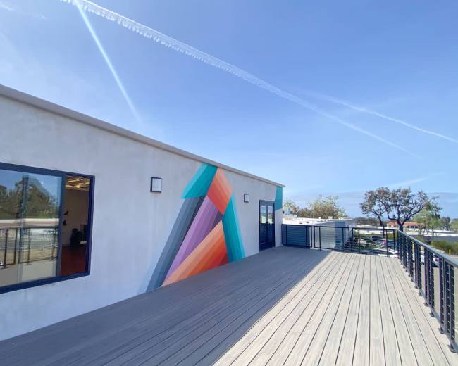 The image shows a modern building with a colorful geometric mural on one wall, surrounded by a sunlit deck and a clear blue sky.