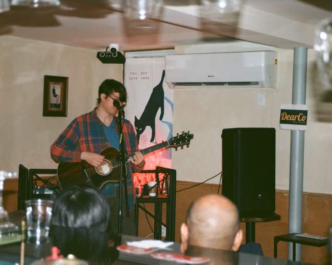 A musician performs on stage with a guitar in a cozy venue, while an audience listens in the foreground.