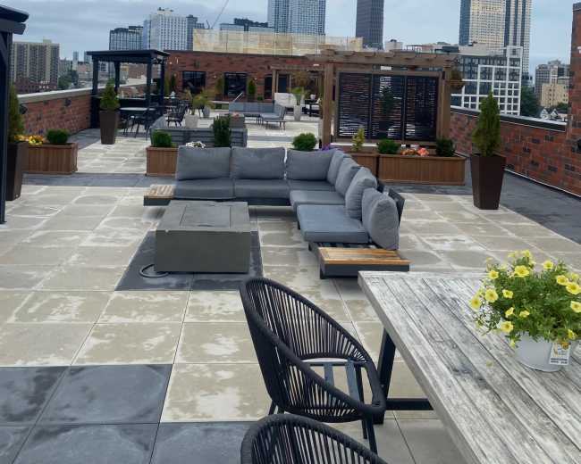 The image shows a rooftop terrace featuring gray couches, wooden dining tables, and various potted plants, with a skyline of tall buildings in the background.