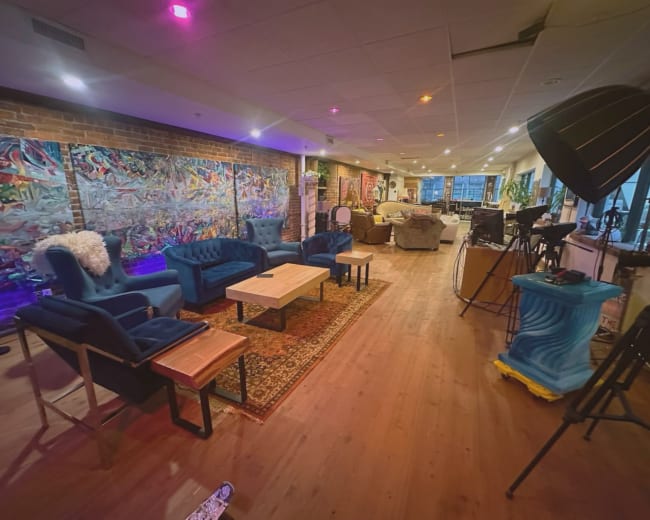The image shows a spacious, modern lounge area with blue armchairs, a wooden table, artistic wall decor, and various seating arrangements in a well-lit environment.