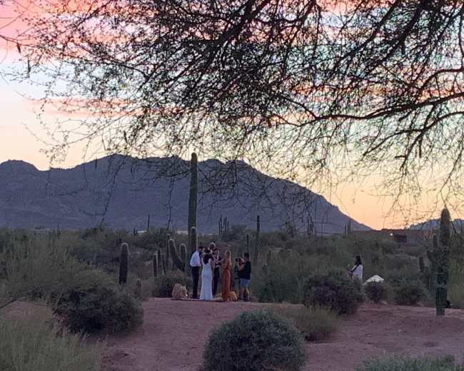 A group of people gathers in a desert landscape at sunset, surrounded by cactus plants and mountains in the distance.