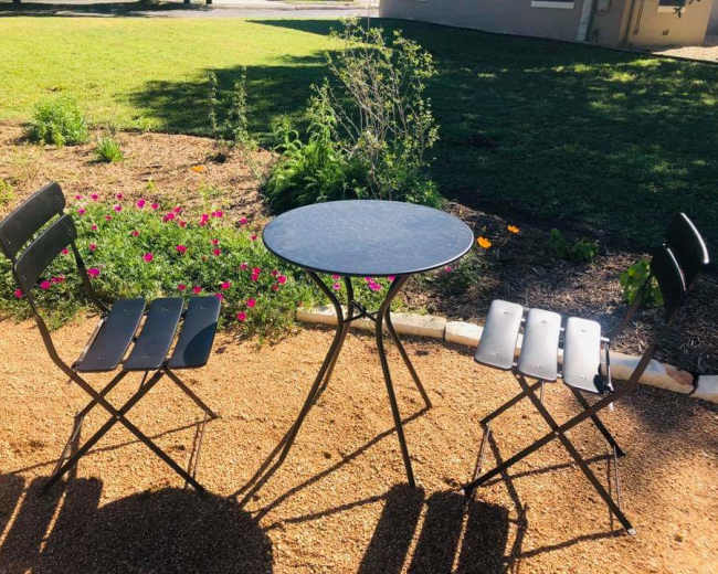 A small round table is positioned between two folding chairs on a gravel patch surrounded by flowers and greenery.