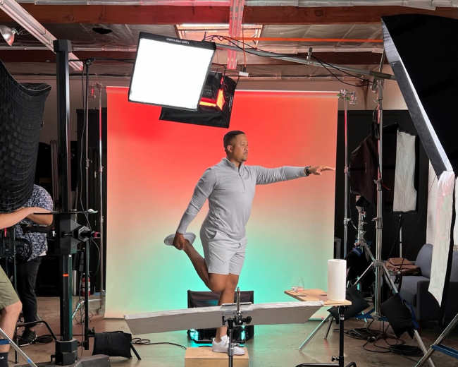 A person in a gray outfit is posing mid-action on a set with a colored backdrop and various studio lighting equipment.