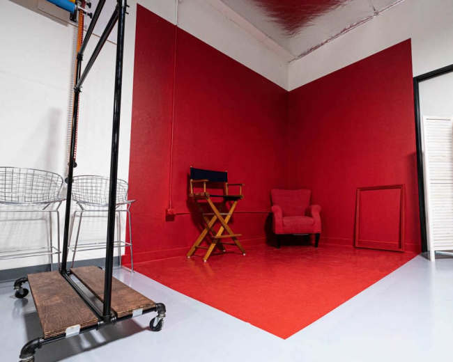 The image shows a small studio corner with a red wall, a chair, a folding screen, and a black clothing rack beside metal stools.