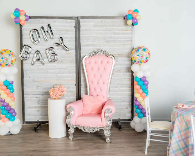 The scene features a decorative area with a pink throne chair, balloon columns, a sign saying "Oh Baby," and a table set with a plaid tablecloth.