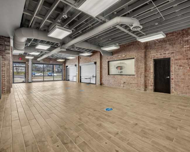 The image shows an empty interior space with wooden flooring, exposed brick walls, and large windows providing natural light.