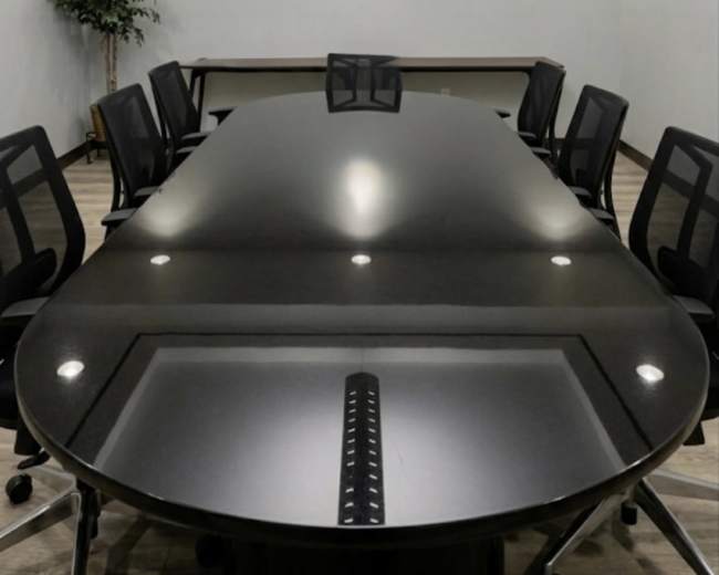 A long, oval conference table surrounded by black rolling chairs is set in a well-lit meeting room with gray walls and a small potted plant in the corner.