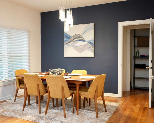 A dining room features a round table with six mustard-colored chairs, a large wall art piece, and a dark blue accent wall.