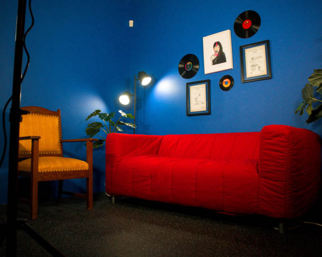 The image shows a bright blue room featuring a red sofa, a wooden chair, and framed records and certificates on the wall, along with a floor lamp illuminating the space.