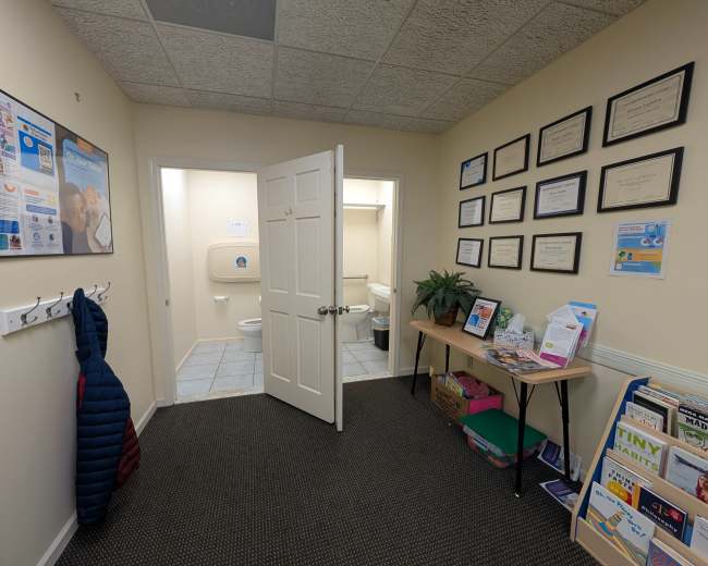 The image shows a small reception area leading to a restroom, with informational materials and framed certificates displayed on the walls.