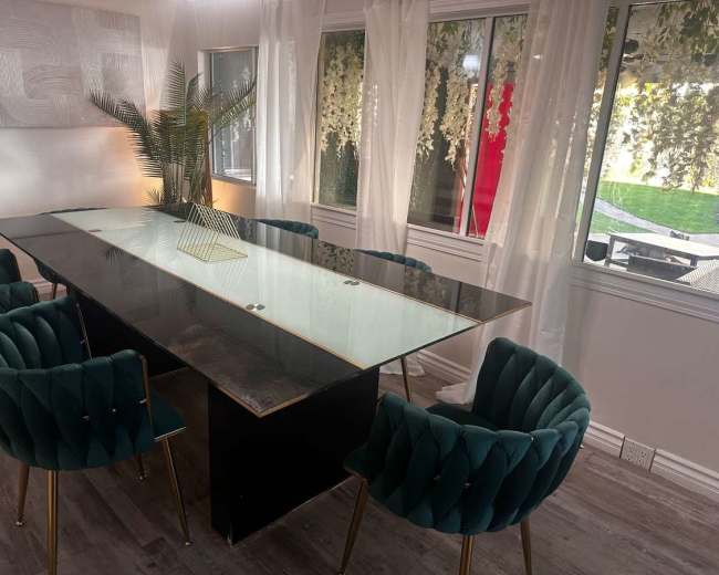 A modern dining area features a sleek glass-top table surrounded by green velvet chairs and large windows letting in natural light.