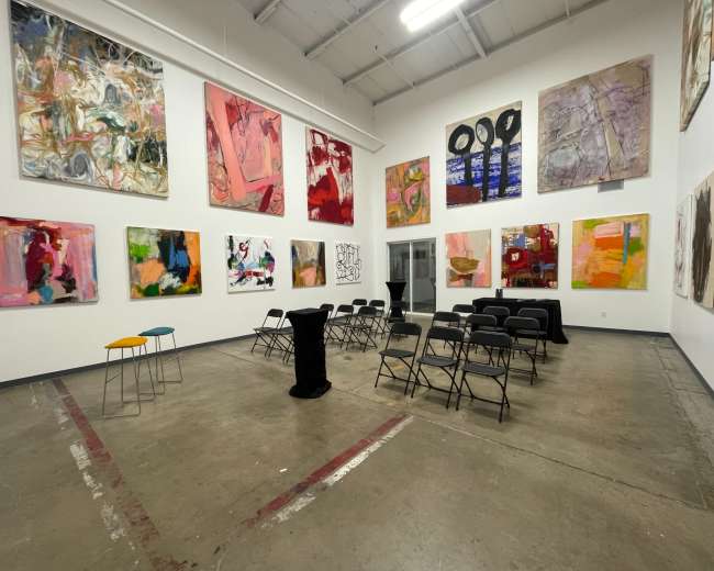 The image shows a modern art gallery with colorful paintings on the walls and a seating area set up with black chairs and small tables.