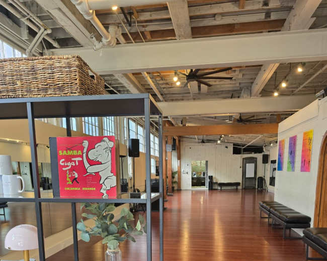 The image shows an expansive, well-lit interior space featuring polished wooden floors, a display shelf with decorative items, and various artworks on the walls.