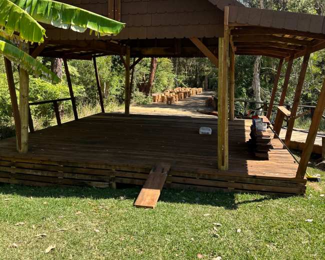 A wooden pavilion with a thatched roof stands on a grassy area surrounded by trees and a pathway lined with timber.