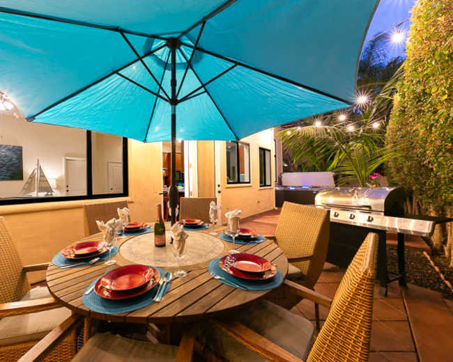 A patio dining setup features a round table with red plates and glasses under a large blue umbrella, accompanied by a grill and surrounded by greenery.