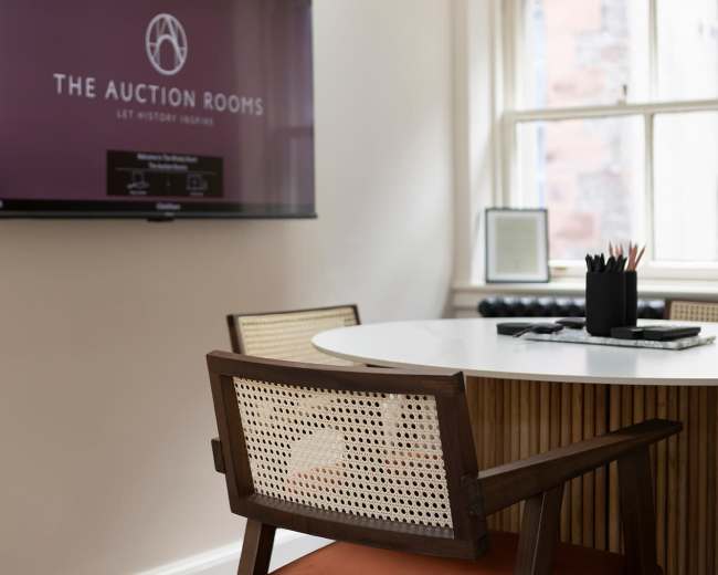 A modern meeting room features a round table, a wooden chair, and a large wall-mounted screen displaying the logo of "The Auction Rooms."