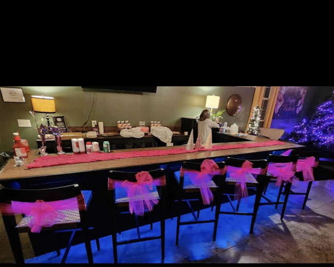 A decorated bar area features pink bows on chairs, a long table runner, and purple lights illuminating a nearby tree.