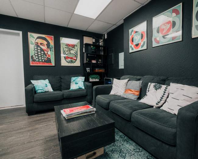 The image shows a stylish waiting room with a dark wall, a grey sofa, decorative pillows, framed artwork, and magazines on a coffee table.