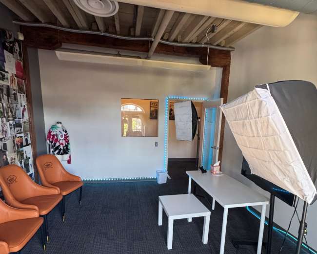 The image shows a spacious room with orange chairs, a white table, and a softbox lighting setup, featuring walls adorned with various images and a visible doorway.
