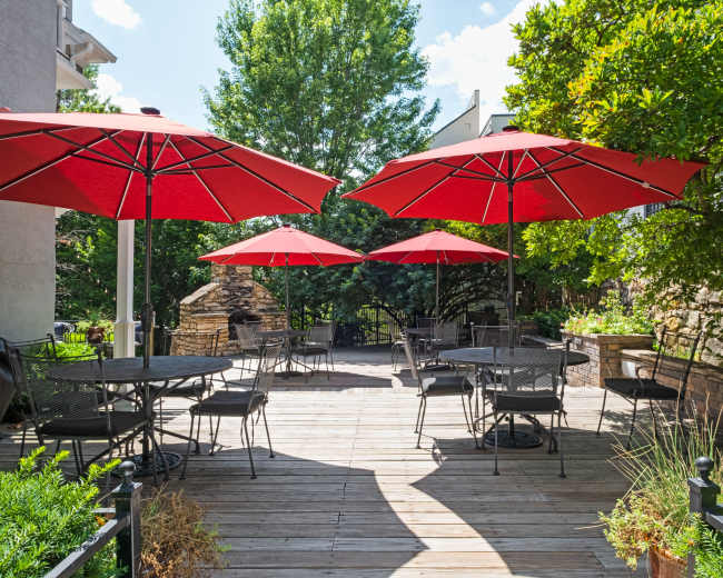 The image shows a wooden deck area with black metal tables and chairs, shaded by large red umbrellas surrounded by greenery.
