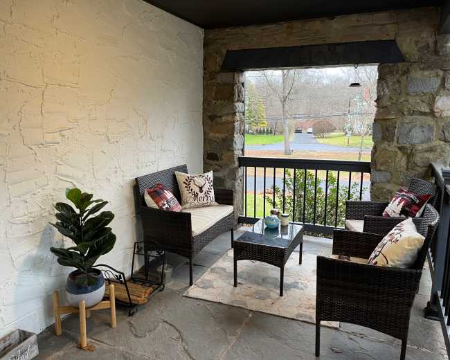 The image shows a cozy porch with wicker furniture, a small coffee table, and a potted plant, set against a stone wall and an outdoor view of a green lawn.