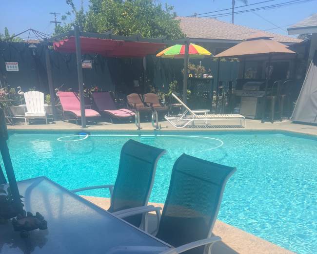 A table with chairs is set near a clear blue swimming pool surrounded by lounging areas and umbrellas.