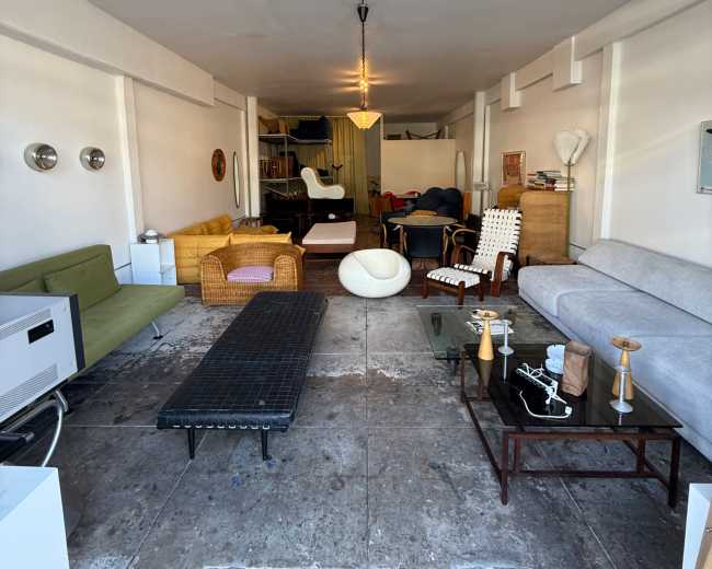 The image shows a long, narrow room furnished with various sofas, chairs, and tables arranged along the walls.