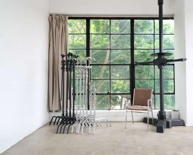 The image shows a minimalist interior with several lighting stands and a single chair beside large windows that overlook greenery outside.