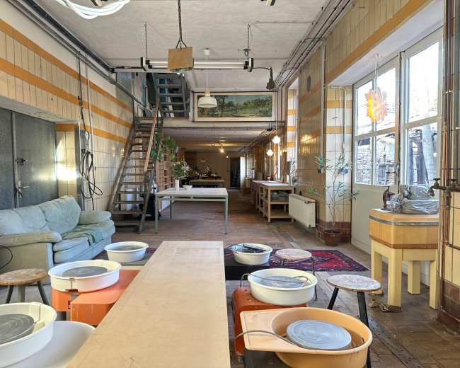 The image shows a spacious, well-lit studio with pottery wheels arranged in a row, a long table in the center, and seating areas along the walls.