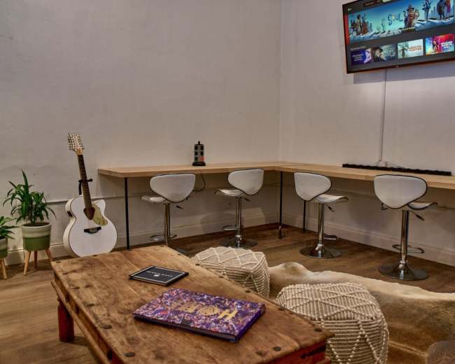The scene features a minimalist lounge area with white bar stools arranged around a long wooden table, a guitar leaning against the wall, and a television mounted above with various shows displayed.