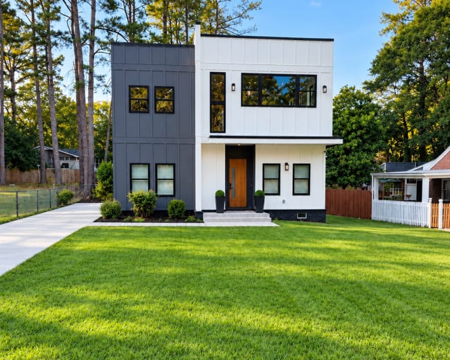 The image features a modern two-story house with a contrasting black and white exterior and a manicured lawn surrounded by trees.