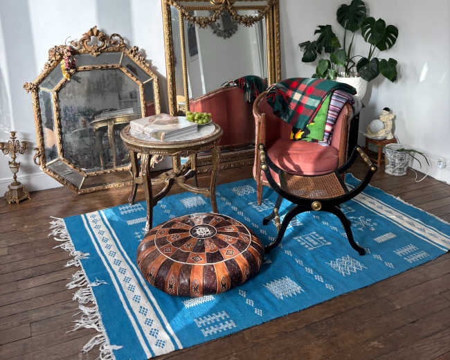 The image shows a cozy corner featuring a vintage mirror, an ornate chair with a colorful blanket, a decorative ottoman, and a potted plant on a blue patterned rug.