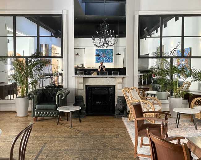 A stylish indoor café or lounge with an arrangement of chairs and small tables, large windows, plants, and a fireplace.