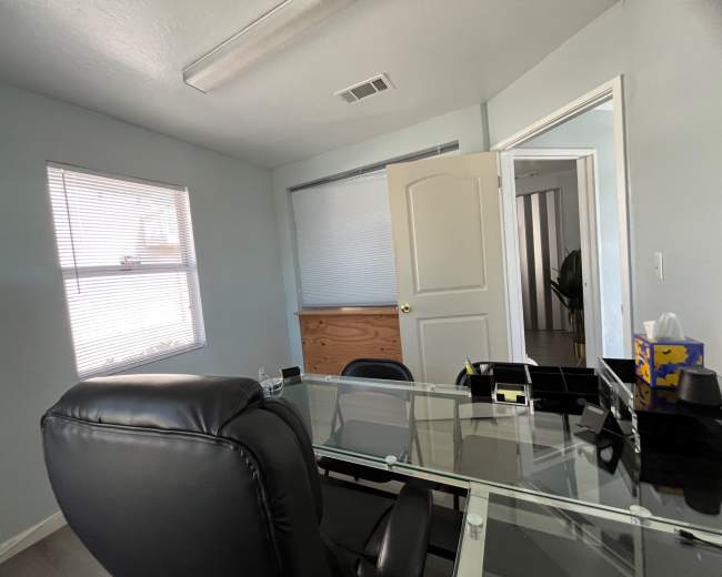 The image shows a small office with a glass desk, black office chairs, and a window, along with an open door leading to another room.